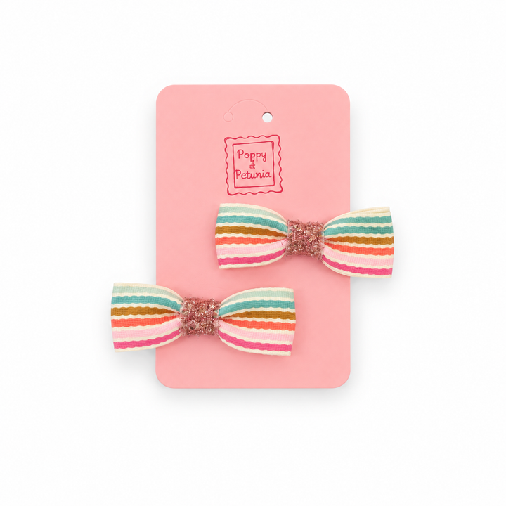 Two rainbow striped mini bow hair clips with rose gold glitter centers on a pink Poppy + Petunia display card.
